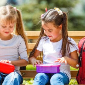 Two young girls sitting on a park bench, opening colourful lunchboxes with backpacks beside them.