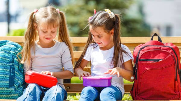Two young girls sitting on a park bench, opening colourful lunchboxes with backpacks beside them.