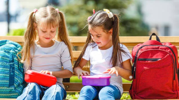 Two young girls sitting on a park bench, opening colourful lunchboxes with backpacks beside them.