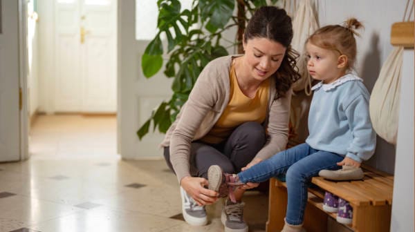 Adult helping a young girl put on her shoes while she sits on a wooden bench in a calm setting.