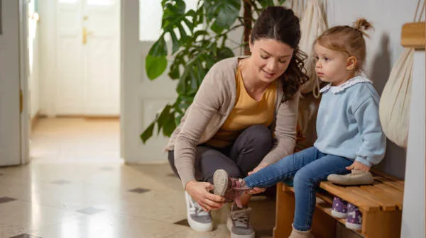 Adult helping a young girl put on her shoes while she sits on a wooden bench in a calm setting.
