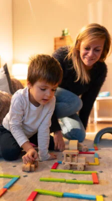 Young boy playing with colorful wooden Montessori toys on the floor while a woman, likely his mother or teacher, watches supportively.
